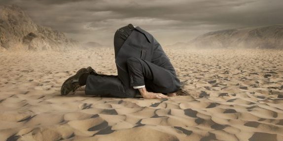 Person wearing suit on knees with head buried in the sand